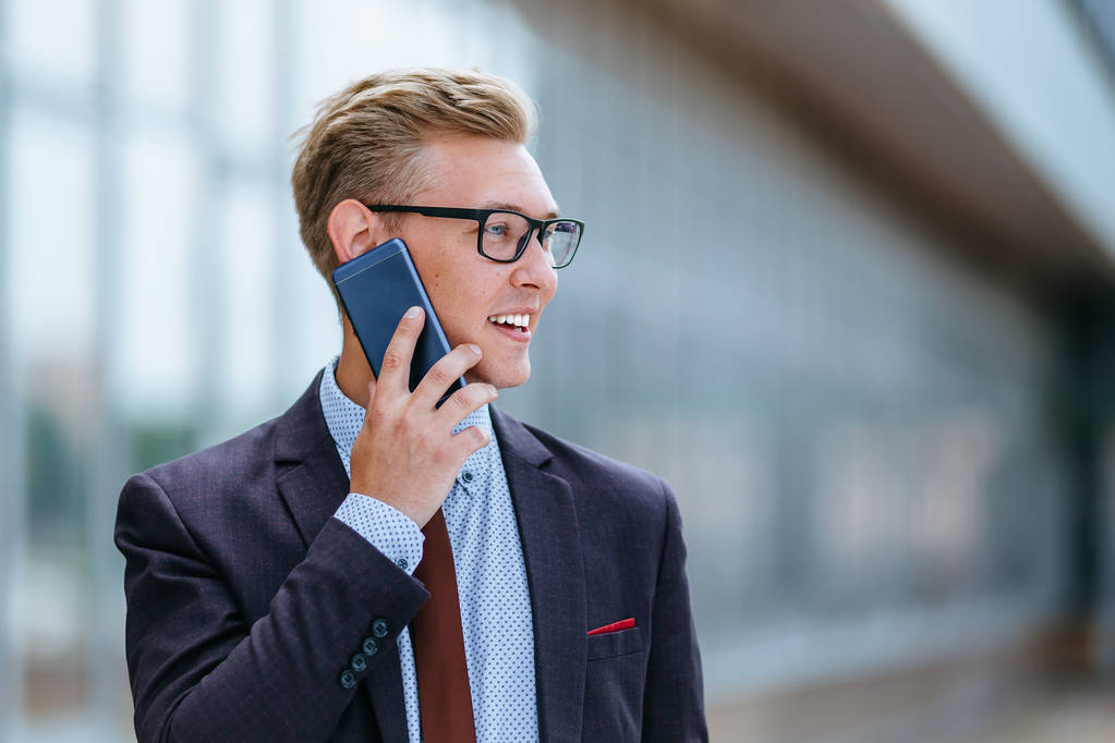 Νέος όμορφος Καυκάσιος επιχειρηματίας με γυαλιά που μιλούν στο κινητό τηλέφωνο μπροστά από το κτίριο του γραφείου. Άντρας εκτελεστικός διευθυντής σε μοντέρνα ρούχα χρησιμοποιώντας smartphone σε εξωτερικούς χώρους. Έννοια της σύγχρονης τεχνολογίας - Φωτογραφία, εικόνα