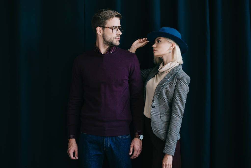 stijlvolle jonge man in glazen en blonde vrouw in hoed kijken naar elkaar in de buurt van gordijn - Foto, afbeelding