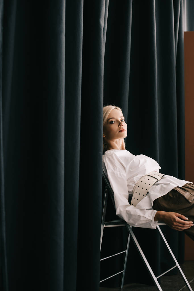 stylische blonde Frau in weißer Bluse sitzt auf Stuhl neben Vorhang  - Foto, Bild