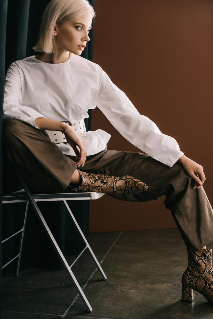 stylish blonde woman in white blouse and boots with snakeskin print sitting on chair near curtain on brown - Photo, Image