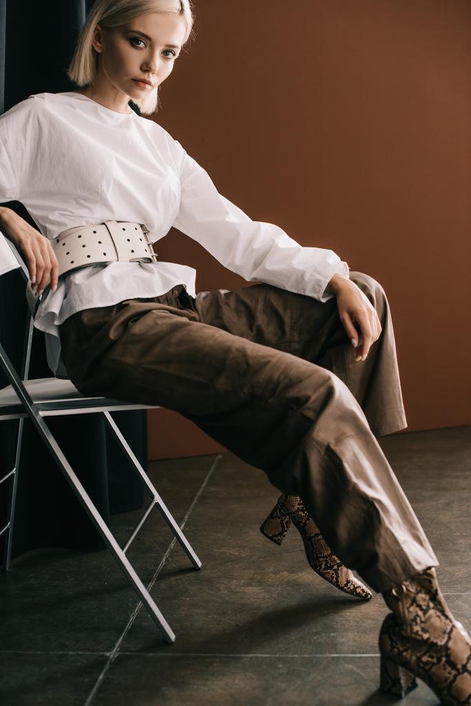 mulher loira elegante em blusa branca e botas com impressão de pele de cobra sentado na cadeira perto de cortina em marrom
 - Foto, Imagem