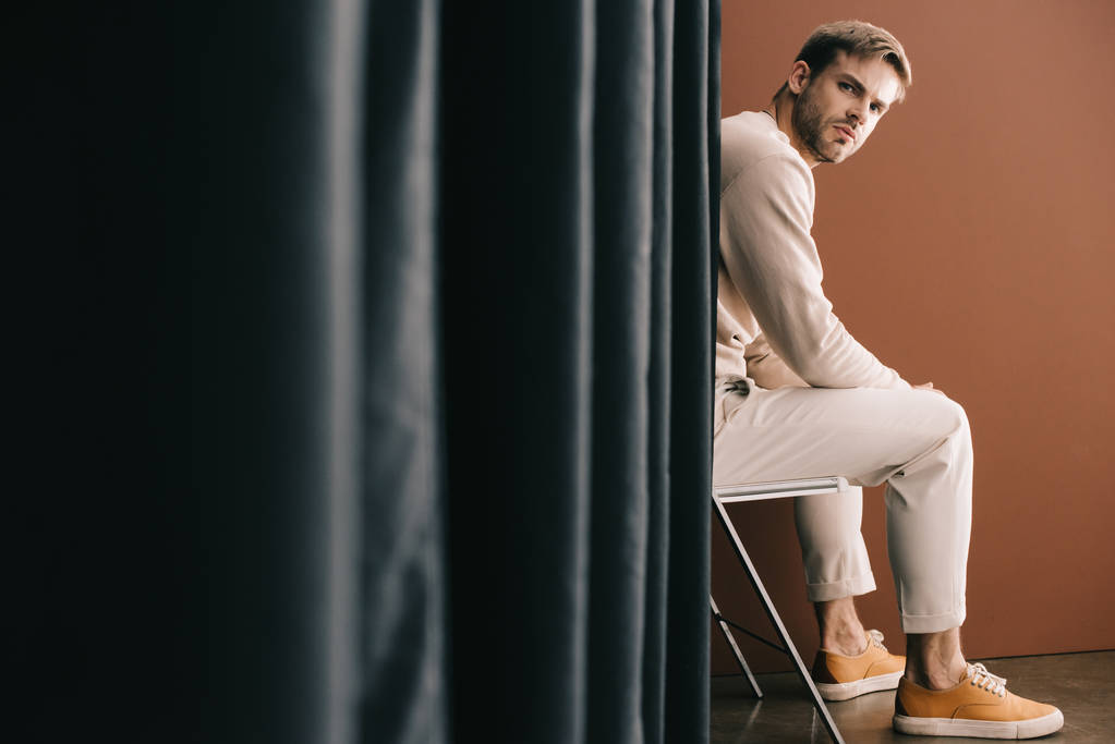 man in casual outfit sitting on chair near curtain on brown - Photo, Image