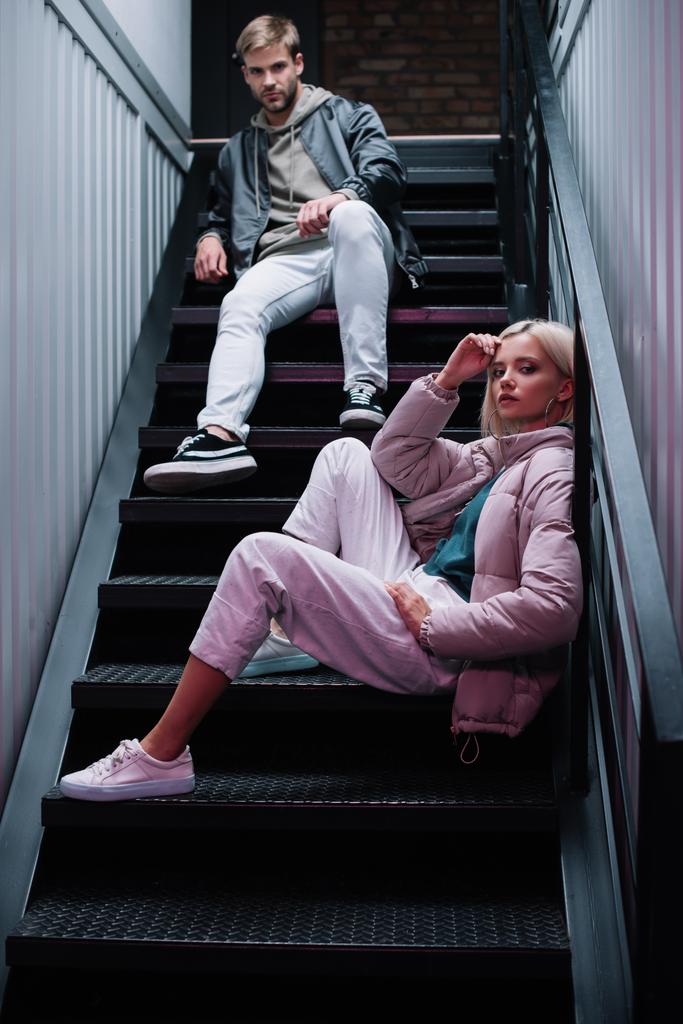 stylish young man and woman in casual autumn outfits sitting on stairs and looking at camera - Photo, Image