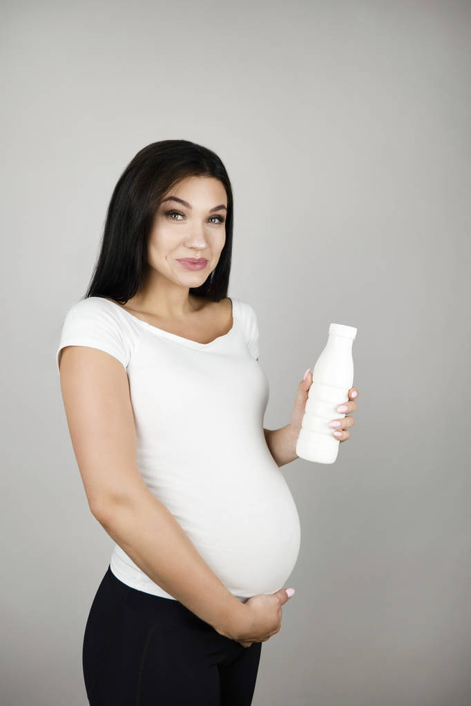 krásná těhotná brunetka, držící láhev s kefírová s úsměvem na izolovaném bílém pozadí - Fotografie, Obrázek