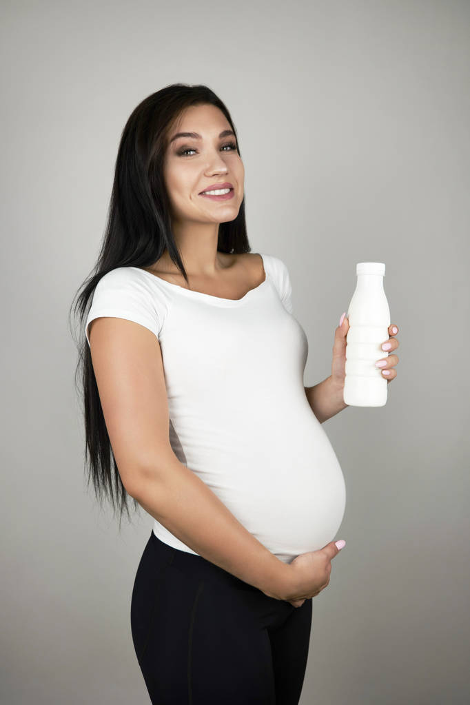hermosa embarazada morena celebración botella con yogur sonriendo sobre aislado blanco fondo
 - Foto, Imagen