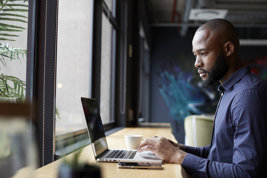 Середній дорослий чорний чоловік креативний сидить біля вікна в офісній соціальній зоні, використовуючи ноутбук, вид збоку
 - Фото, зображення