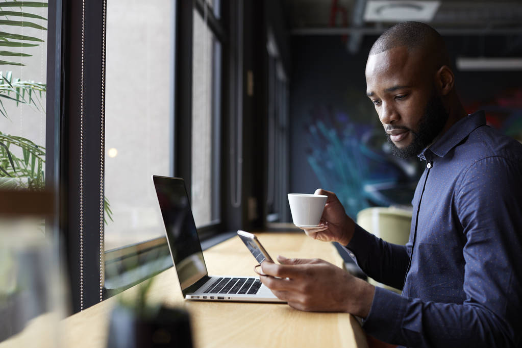 Mid dorosły czarny mężczyzna twórczy siedzi przez okno o kawę, za pomocą laptopa i smartfona, widok z boku - Zdjęcie, obraz