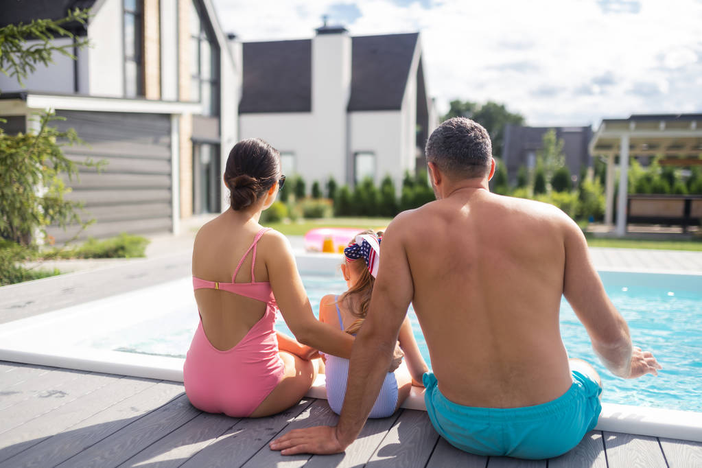 Семья в купальниках, сидящая у бассейна рядом с частным домом
 - Фото, изображение