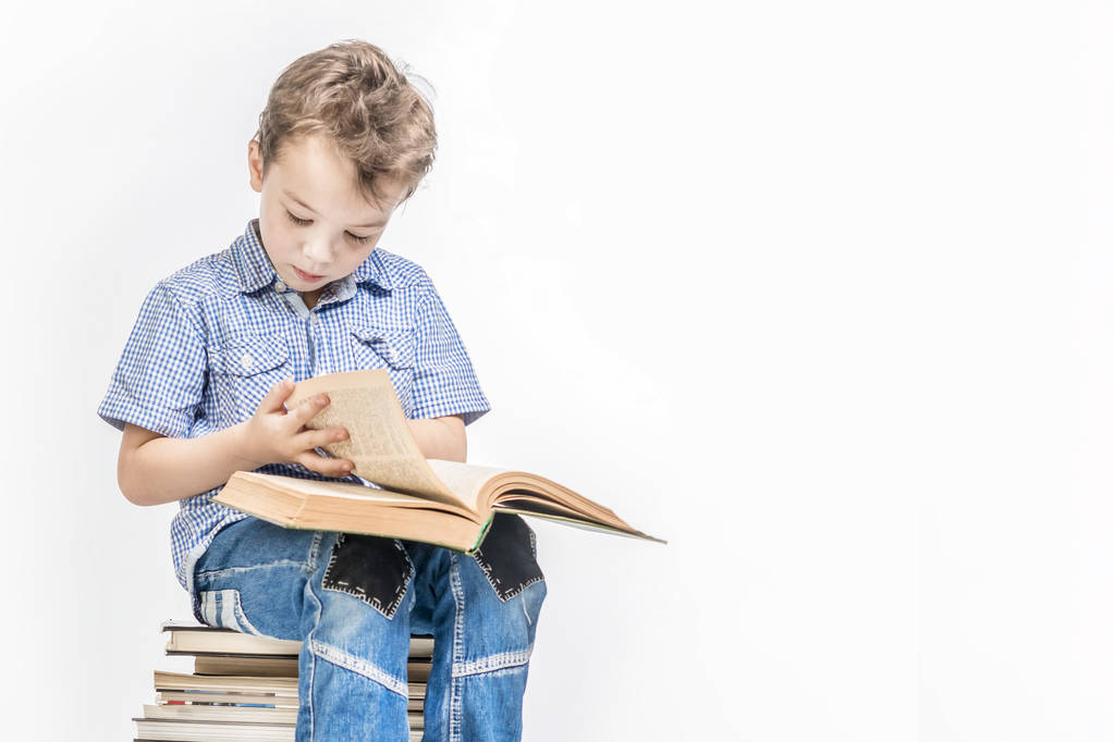 Ένα όμορφο αγόρι κάθεται σε μια στοίβα από βιβλία και διαβάζει ένα βιβλίο στο - Φωτογραφία, εικόνα