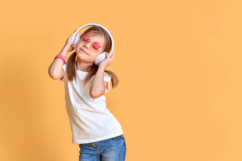 Дівчина слухає музику в навушниках на жовтому фоні. Мила дитина насолоджується щасливою танцювальною музикою, близькими очима і посмішкою позує на стіні студії
 - Фото, зображення