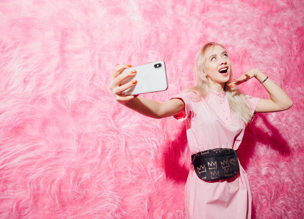 Яскрава молода дівчина-блогер, одягнена в модну рожеву сукню, бере селфі на свій смартфон на фоні рожевої хутряної стіни в шоу-румі
 - Фото, зображення