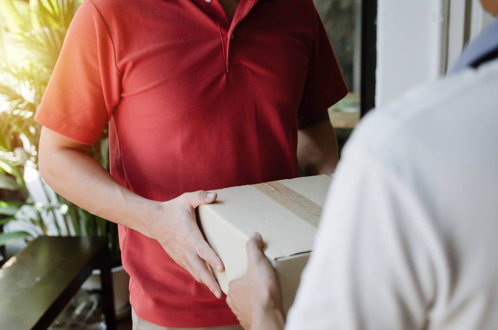hand of young delivery service man in red uniform with man customer receiving parcel post box from courier at home, cargo shipping, express delivery service, online shopping and logistics concept - Photo, Image