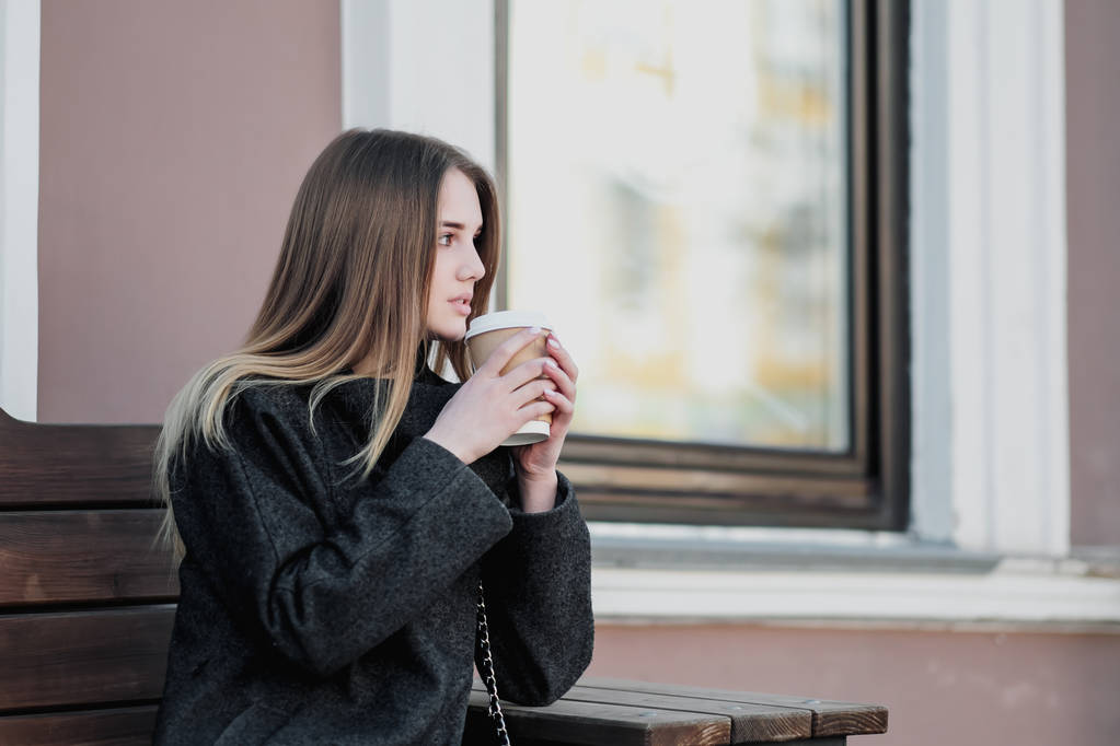 Menina milenar bonita nova em um casaco está sentado em um banco de outdor com uma xícara de papel de café. Outono ou dia de primavera, tempo frio
. - Foto, Imagem