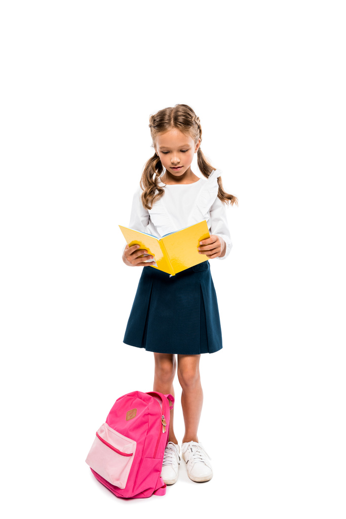 Śliczne dziecko czytanie książki i stojący w pobliżu różowy plecak na białym tle  - Zdjęcie, obraz