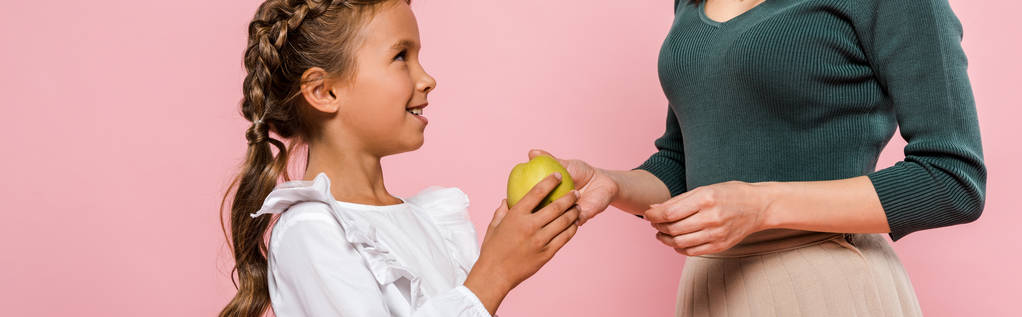 Panoramaaufnahme einer Mutter, die ihrer Tochter einen leckeren Apfel gibt, isoliert auf rosa  - Foto, Bild