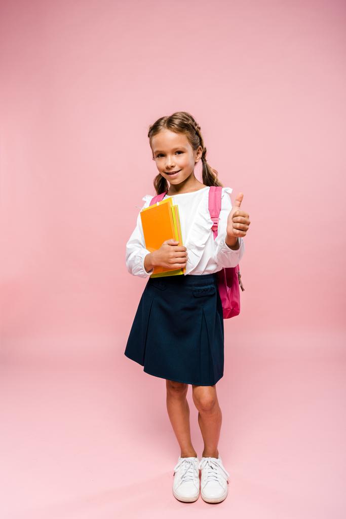 glückliches Kind, das Bücher hält, während es mit dem Rucksack steht und den Daumen auf rosa zeigt  - Foto, Bild