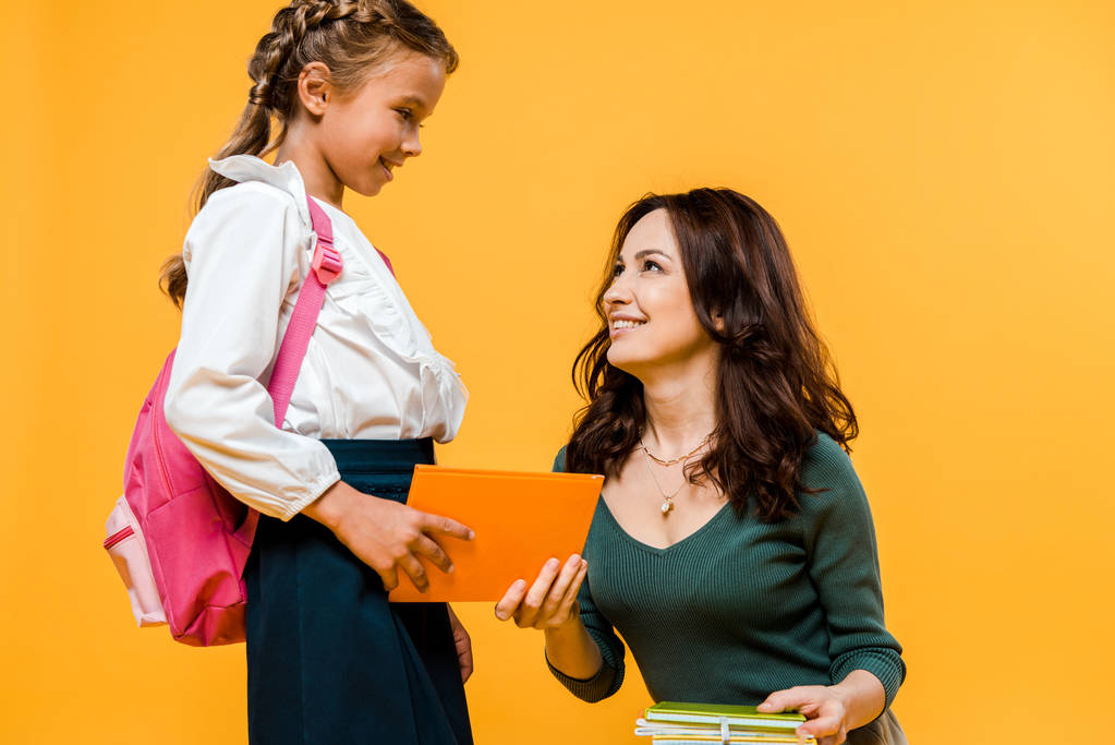 ελκυστική μητέρα δίνοντας βιβλίο σε ευτυχισμένο μαθήτρια απομονωμένη στο πορτοκαλί  - Φωτογραφία, εικόνα