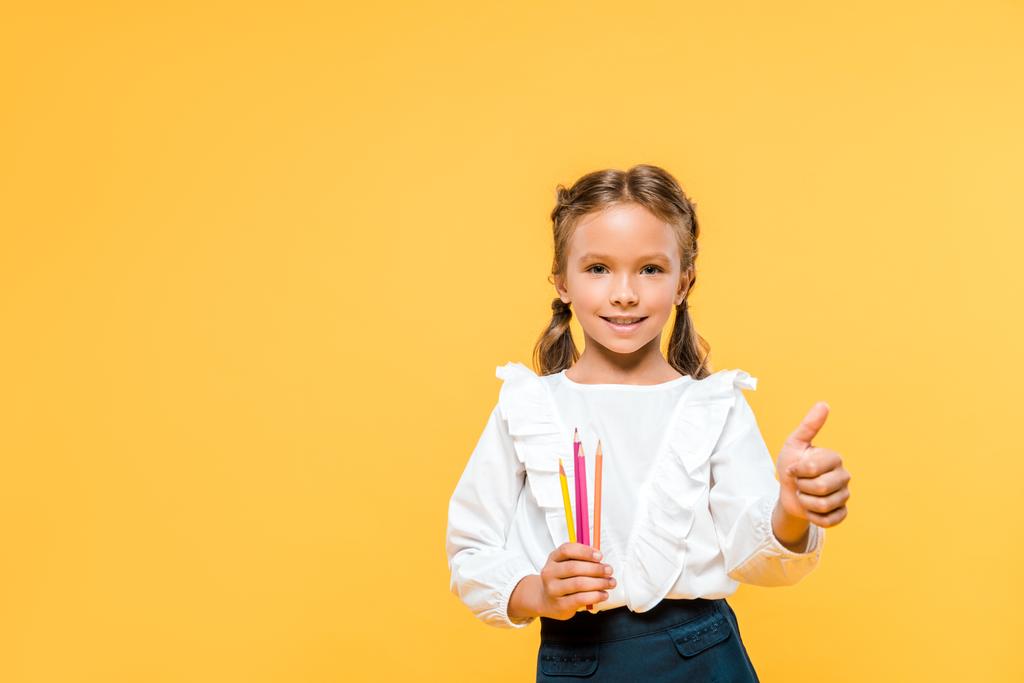 счастливый школьник держит цветные карандаши и показывает большой палец вверх изолированы на оранжевый
  - Фото, изображение