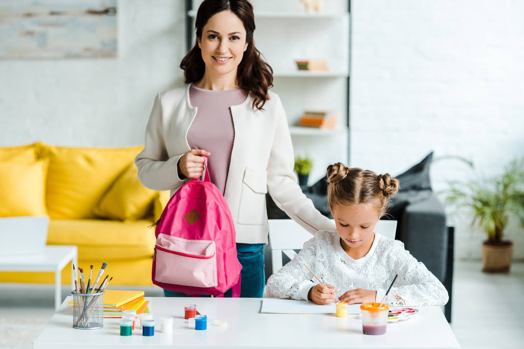feliz mãe segurando mochila perto de bonito filha pintura em casa
  - Foto, Imagem