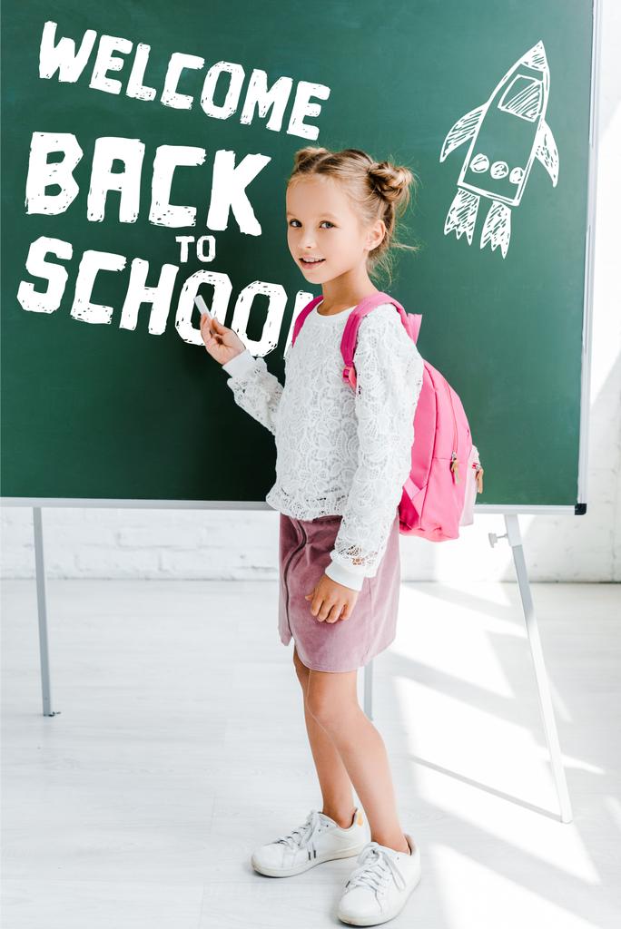 Милая школьница, стоящая с рюкзаком и держа мел рядом с Добро пожаловать обратно в школу буквы на зеленой доске
  - Фото, изображение