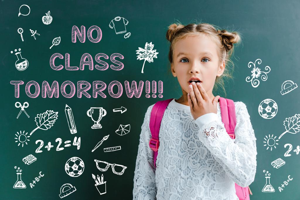 surprised schoolgirl covering mouth near chalkboard with no class tomorrow lettering on green  - Photo, Image