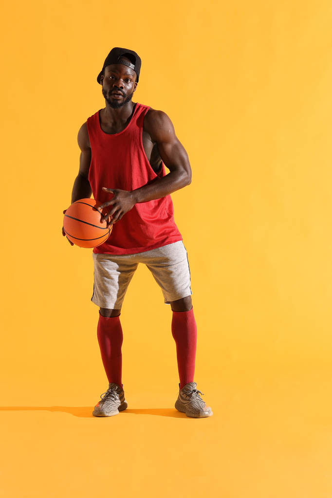 Full length portrait of black man in red shirt, black cap and grey shorts playing basketball. Studio, yellow background - Photo, Image