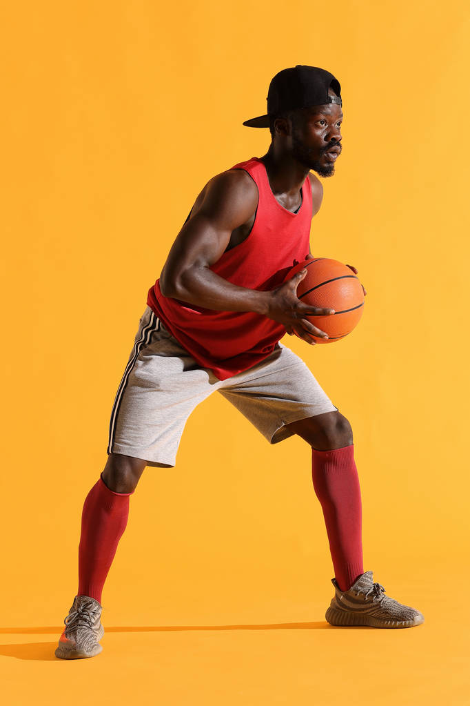 Ολόσωμο πορτρέτο μαύρου με κόκκινο πουκάμισο, μαύρο καπάκι και γκρι σορτς παίζοντας μπάσκετ. Στούντιο, κίτρινο φόντο - Φωτογραφία, εικόνα