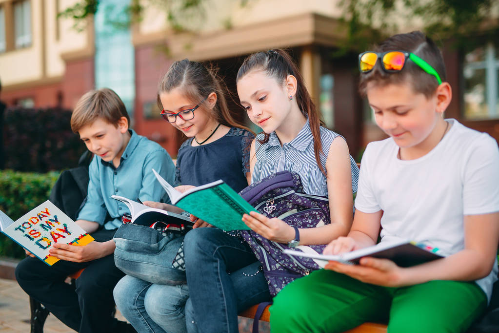 Boldog iskolatársak portréja. Iskolatársak ülnek könyvek egy fa padon a városi parkban, és tanul a napsütéses napon. - Fotó, kép