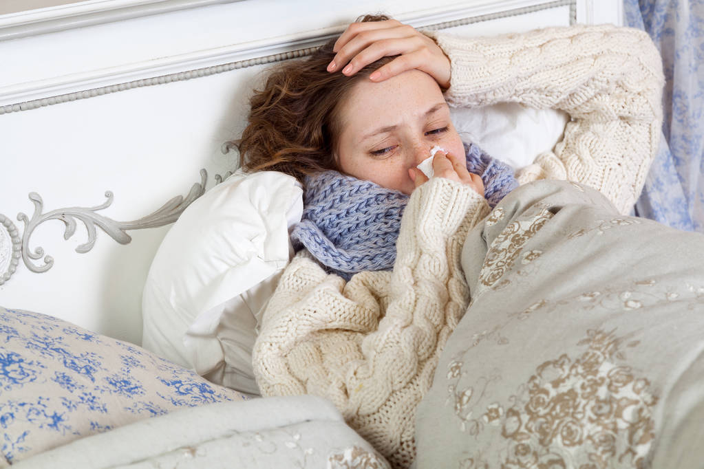 Nuori nainen valkoisessa villapaita ja sininen huivi puhdistus nenänsä ja tilalla tuskallinen pää tunteessaan kylmä sairas ja lepää kotona sängyssä
  - Valokuva, kuva