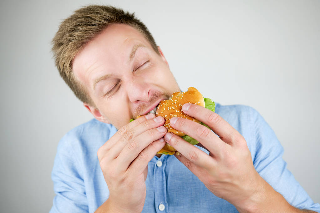 Молодой красивый мужчина кусает свежий вкусный бургер из фаст-фуда ресторан выглядит голодным есть на изолированном белом фоне
 - Фото, изображение