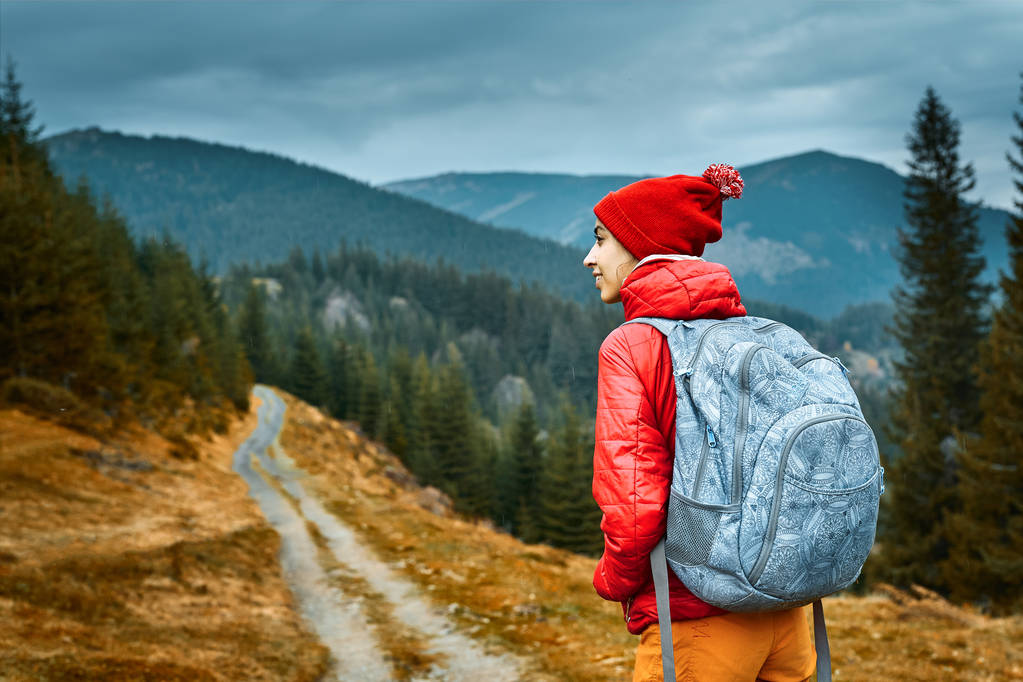 mujer excursionista con mochila, con chaqueta roja y pantalones anaranjados, de pie sobre el fondo de las montañas
 - Foto, Imagen