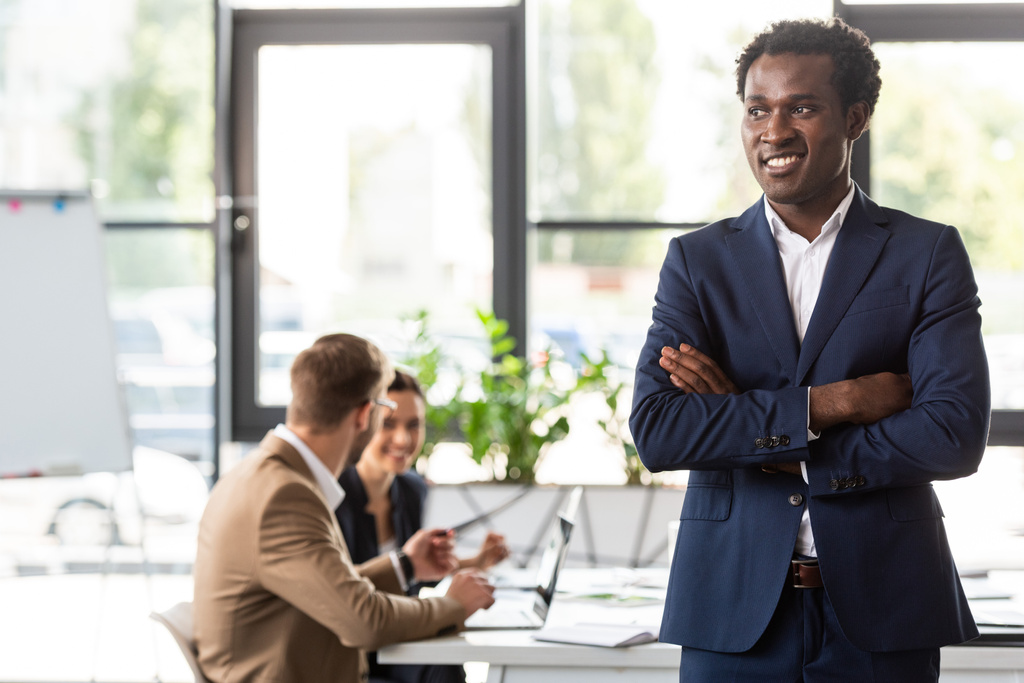 uśmiechnięty African American biznesmen w formalne nosić stojący z skrzyżowanymi ramionami przed kolegami w biurze - Zdjęcie, obraz