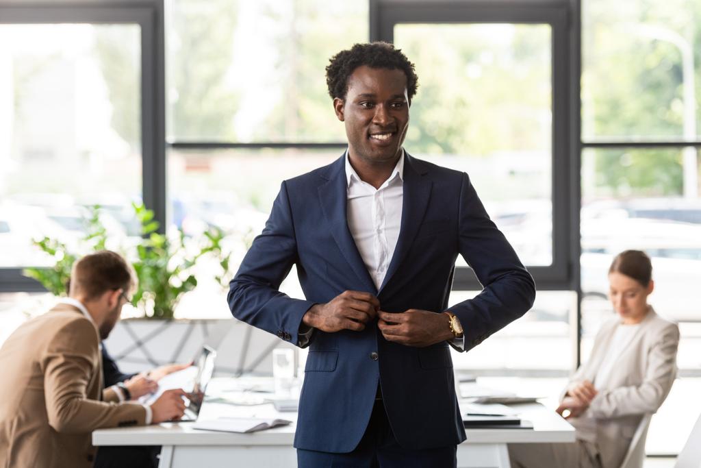 glimlachend Afro-Amerikaanse zakenman in formele slijtage staande voor collega's in Office - Foto, afbeelding