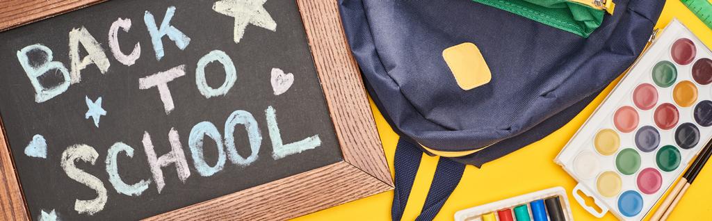 Πανοραμική βολή μαυροπίνακα σε ξύλινο πλαίσιο με πίσω στο σχολείο επιγραφή κοντά σε μπλε σχολική τσάντα και υδατογραφικά χρώματα που έχουν οριστεί σε κίτρινο φόντο - Φωτογραφία, εικόνα