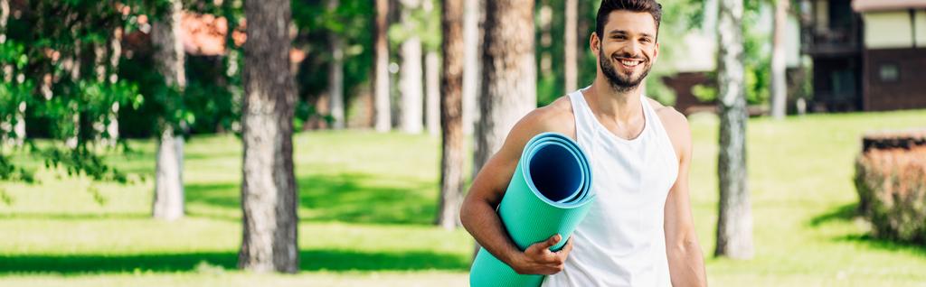 panoramatický záběr šťastného atletického sportovce, který drží fitness rohožku v parku  - Fotografie, Obrázek