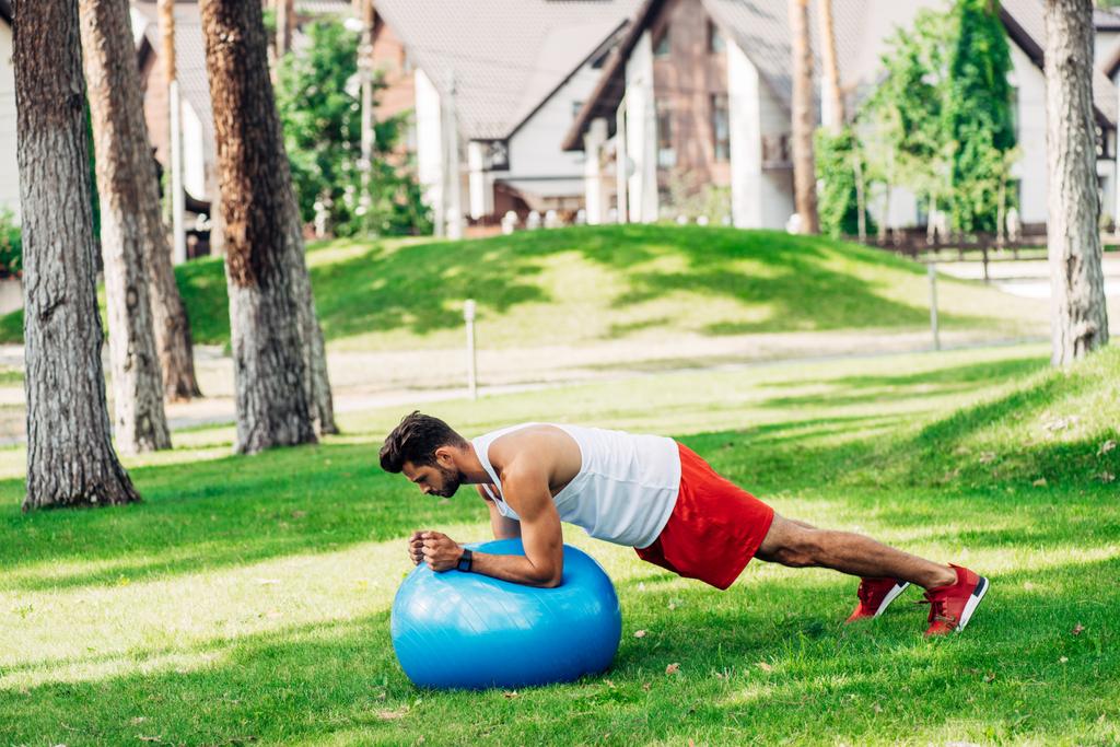 Brodaty mężczyzna w odzieży sportowej wykonywania na niebieskim piłkę fitness w parku  - Zdjęcie, obraz