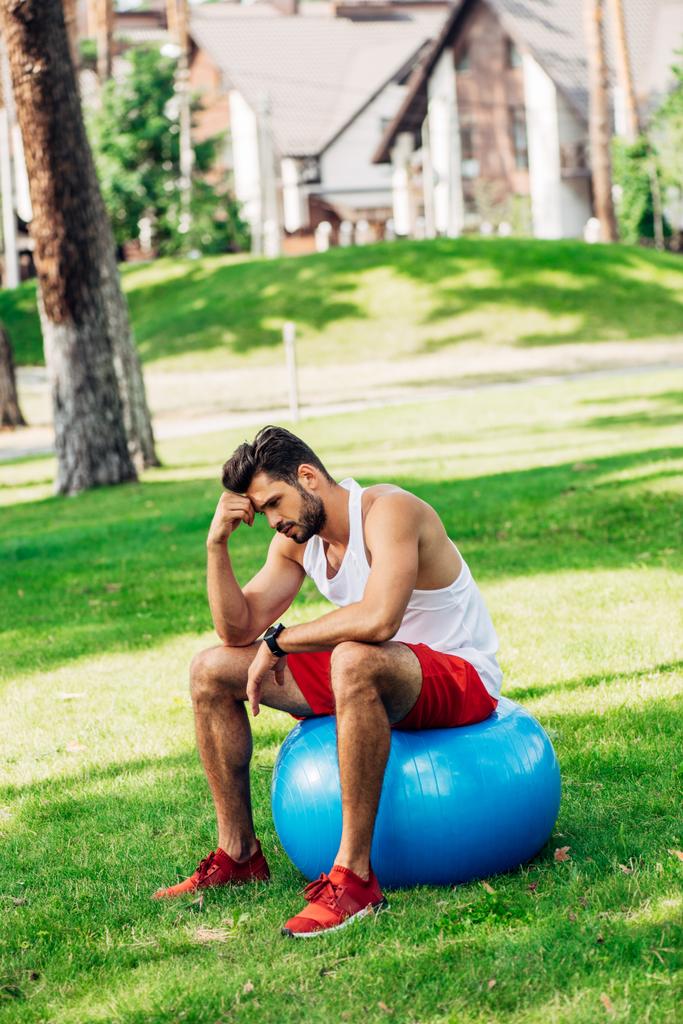 zdenerwowany sportowcem siedzi na niebieskim piłkę fitness w parku  - Zdjęcie, obraz