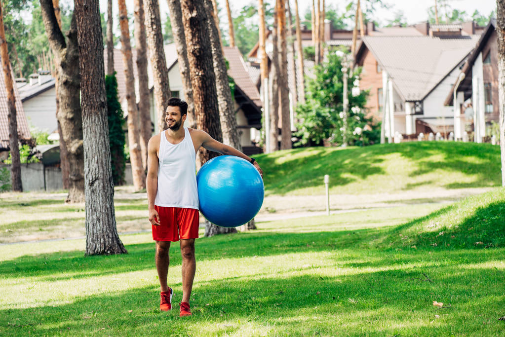 glücklicher Sportler, der auf grünem Gras geht und Fitnessball hält  - Foto, Bild