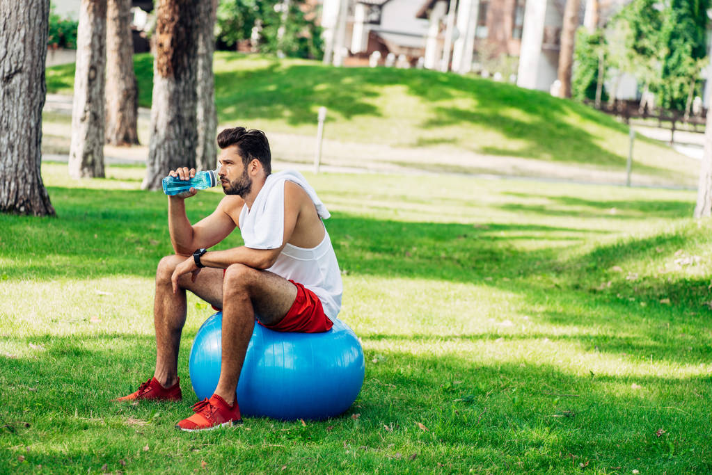 bärtiger Sportler trinkt Wasser aus Sportflasche, während er auf Fitnessball sitzt - Foto, Bild