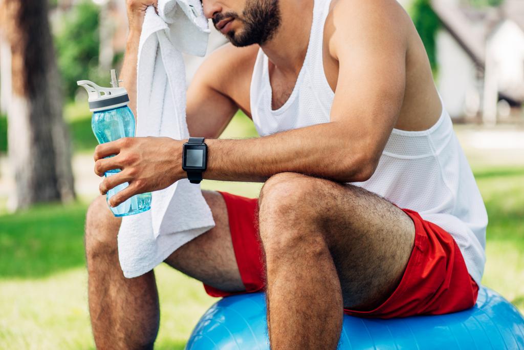 abgeschnittene Ansicht eines bärtigen Mannes mit Sportflasche, während er auf einem Fitnessball sitzt und sich den Schweiß abwischt - Foto, Bild