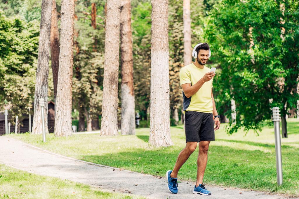 ευτυχισμένος μουσάτος άνθρωπος χρησιμοποιώντας smartphone και ακούγοντας μουσική στα ακουστικά, ενώ το περπάτημα στο πάρκο  - Φωτογραφία, εικόνα