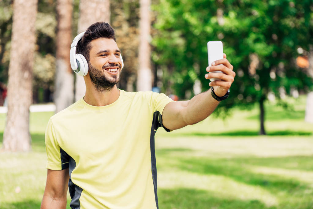 счастливый бородатый мужчина делает селфи, держа смартфон и слушая музыку в наушниках
 - Фото, изображение