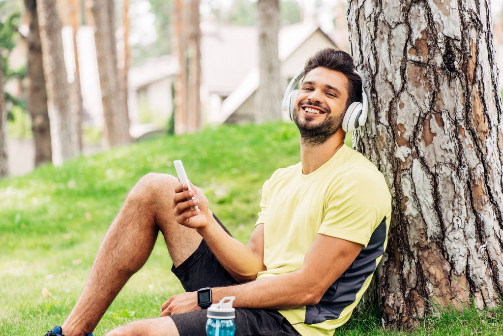 счастливый мужчина в наушниках сидит возле дерева и держит смартфон
  - Фото, изображение