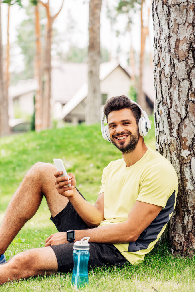 happy man in headphones sitting near tree trunk and holding smartphone  - Photo, Image