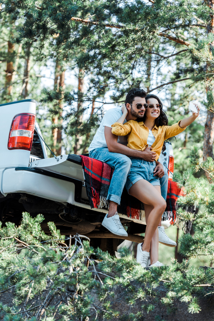 fröhliches Mädchen spricht Selfie mit Mann in der Nähe von Auto und Bäumen  - Foto, Bild
