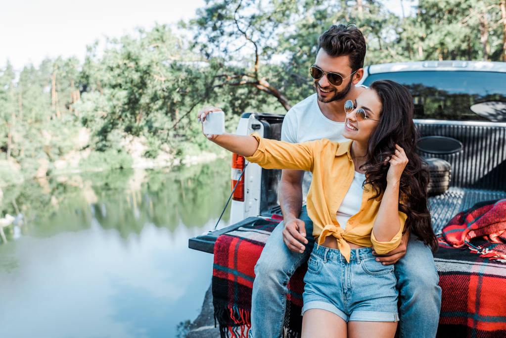 iloinen tyttö aurinkolasit puhuminen selfie miehen kanssa lähellä autoa ja järveä
 - Valokuva, kuva