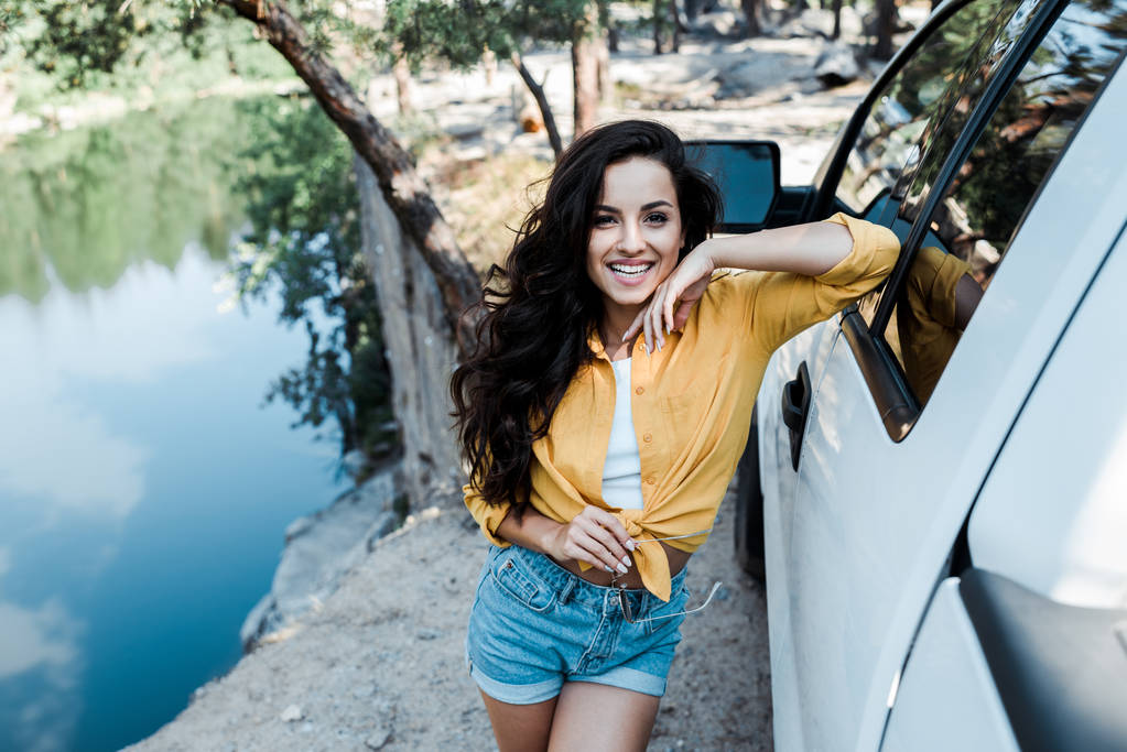 selective focus of happy girl smiling while standing near car in woods  - Photo, Image