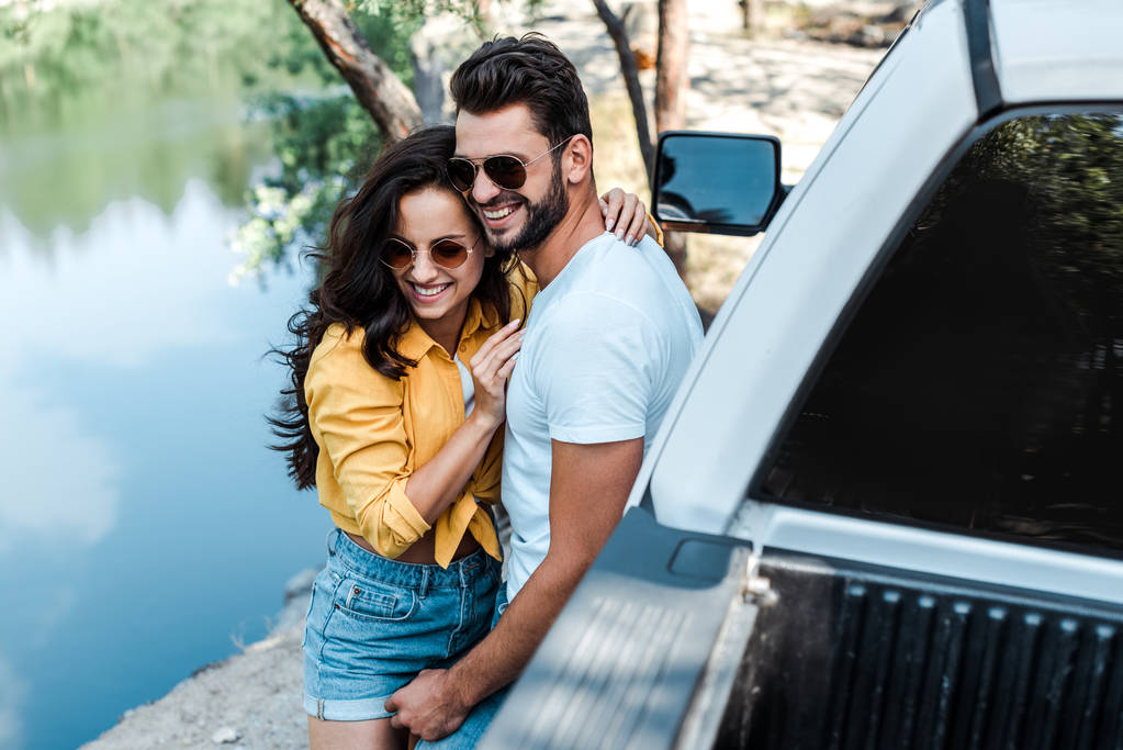 enfoque selectivo de hombre barbudo abrazando a chica atractiva en gafas de sol cerca del coche
  - Foto, imagen