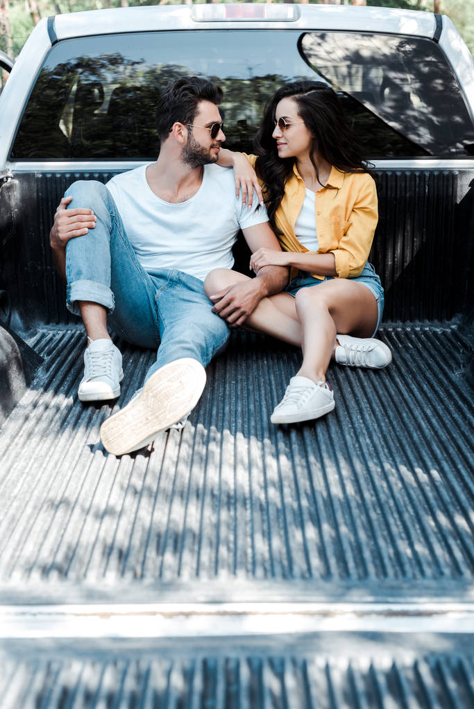 cheerful man in sunglasses sitting in car trunk and looking at girl  - Photo, Image