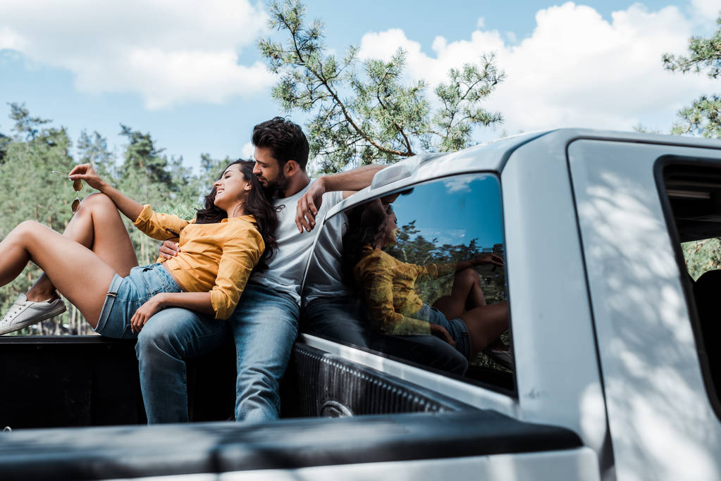 Tiefansicht eines gutaussehenden bärtigen Mannes, der auf einem Auto sitzt und eine glückliche junge Frau ansieht  - Foto, Bild